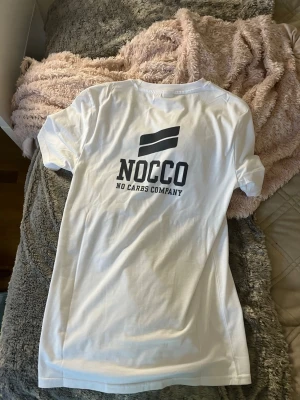 Vit NOCCO t-shirt med tryck - Säljer en vit t-shirt från NOCCO med stort svart tryck på ryggen där det står 'NOCCO No Carbs Company' och en grafisk logga ovanför. T-shirten har korta ärmar och är i mjukt bomullsmaterial. Perfekt för dig som gillar sportiga och stilrena plagg. Det enda fel med den är att nocco märker på bröstet står spegelvänt men inget man tänker på så mycket. Aldrig använd 