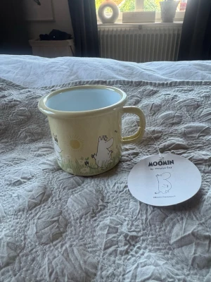 Moomin emaljmugg från Muurla - Söt emaljmugg med Muminmotiv från Muurla. Muggen är ljusgul med illustrationer av Mumintrollet och andra figurer i grönt gräs och sol. Perfekt för te, kaffe eller varm choklad. Lätt och tålig, passar både hemma och på utflykt. Rymmer 2,5 dl är är helt oanvänd