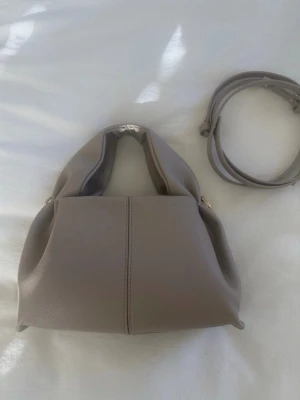 Polene Beige handväska  - Stilren beige handväska i mjukt konstläder med avtagbar axelrem och guldfärgade metalldetaljer. Väskan har en modern, boxig form och dubbla handtag för olika bärstilar. Perfekt för dig som gillar minimalistisk design och neutrala färger.