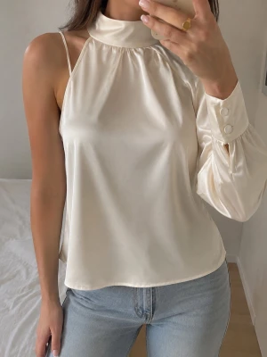 Krämvit satinblus med asymmetrisk ärm - Snygg krämvit blus i satin med hög krage och unik asymmetrisk design. Ena sidan har smalt axelband och ärmlös look, medan andra sidan har lång puffärm med bred manschett och knappar. Perfekt för dig som vill sticka ut med en trendig topp.