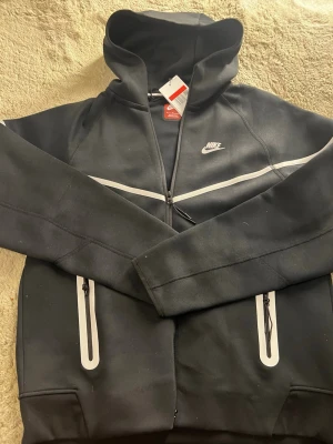 Svart Nike hoodie med dragkedja - Snygg svart hoodie från Nike med vit logga på bröstet och vita detaljer längs dragkedjan och fickorna. Hoodien har hel dragkedja, två fickor med dragkedja och en klassisk huva. Perfekt för en sportig och avslappnad stil.