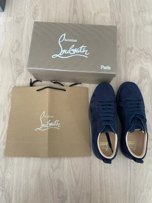 Mörkblåa Christian Louboutin sneakers - Snygga mörkblåa sneakers från Christian Louboutin i mocka med klassisk röd sula och guldfärgad logga på insidan. Skorna har rund tå, matchande blå skosnören och låg profil som ger en clean och exklusiv vibe.