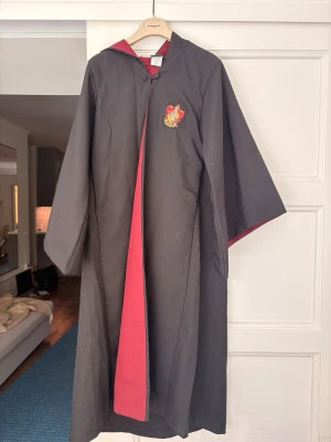 Gryffindor mantel Harry Potter cosplay - Snygg Gryffindor-mantel från Harry Potter med huva och röd insida. Gryffindor-emblem på bröstet. Manteln är svart med röda detaljer och har vida ärmar. Perfekt för cosplay eller maskerad. Tillverkad i polyester.