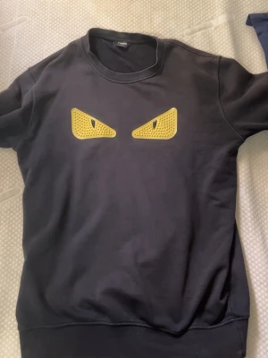 Svart Fendi sweatshirt med gula ögon - Cool svart sweatshirt från Fendi med ikoniska gula ögon på bröstet. Tröjan har rund halsringning och ribbade muddar vid ärmslut och nederkant. Perfekt för dig som vill sticka ut med en unik designertröja.