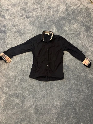 Svart skjorta från Burberry London - Svart skjorta från Burberry London med klassiskt rutigt mönster på insidan av krage och ärmslut. Skjortan har knappar framtill och långärmad design. Perfekt för dig som gillar stilrena plagg med exklusiva detaljer.