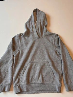 Grå hoodie med gymnastiktryck storlek 140 - Grå hoodie med huva och magficka. På ryggen finns ett rosa tryck med texten 'GYMNASTICS MY PASSION' och en gymnast i siluett. Tillverkad i mjukt sweatshirtmaterial, perfekt för dig som älskar gymnastik och vill ha en chill stil.