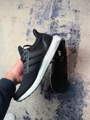 Svarta Adidas Ultraboost sneakers - Säljer ett par svarta Adidas Ultraboost sneakers med stickad ovandel och vita Boost-mellansulor. Skorna har svarta plastdetaljer på sidorna och Continental-gummisula undertill för extra grepp. Perfekta för dig som gillar stilrena och bekväma sneakers.