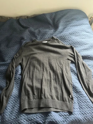 Mörkgrå stickad tröja Jack & Jones - Mörkgrå stickad tröja från Jack & Jones med rund halsringning och ribbade muddar vid ärmslut och nederkant. Enkel och stilren design som passar till det mesta. Perfekt för lager på lager och snygg till jeans.