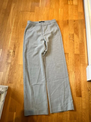Grå kostymbyxor från Gina Tricot - Snygga grå kostymbyxor från Gina Tricot med raka ben och diskret rutmönster. Byxorna har hällor för skärp och klassisk midja. Perfekta för en stilren look och passar till många olika outfits.