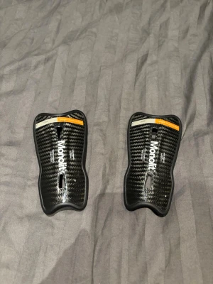 Svarta benskydd från Monolit - Säljer ett par svarta benskydd från Monolit med kolfibermönster och vita detaljer. De har en ergonomisk form och orange markering upptill. Perfekta för fotboll eller andra sporter där skydd behövs. Använda en gång. OBS fodralet finns ej. Pris kan diskuteras 