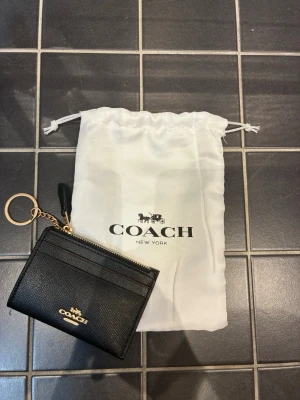 Svart Coach korthållare med nyckelring - Snygg svart korthållare från Coach i läder med guldfärgade detaljer och nyckelring. Framsidan har Coach-logga i guld och flera kortfack, baksidan har ett genomskinligt fack för ID eller busskort. Kommer med vit dustbag med Coach-tryck.