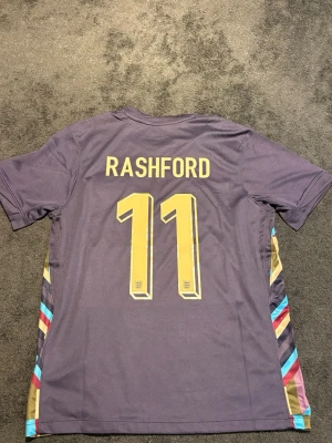 England Rashford matchtröja Nike - Snygg England matchtröja från Nike med Rashford och nummer 11 i gult på ryggen. Tröjan har färgglada detaljer i blått, rött och guld längs sidorna samt Englands landslagsemblem på bröstet. Perfekt för fotbollsfans som vill sticka ut på läktaren eller planen.