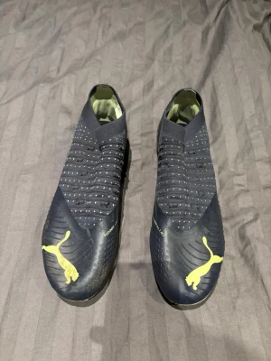 Puma blå fotbollsskor utan snörning - Säljer ett par mörkblå fotbollsskor från Puma med gula detaljer och logga. Skorna har en strumpliknande passform utan snörning och mönstrad ovandel för extra grepp. Sulan är gul med svarta dobbar, perfekt för dig som vill sticka ut på planen. OBS inga snören tillkommer