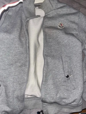 Grå hoodie från Moncler - Snygg grå hoodie från Moncler med dragkedja och ikonisk logga på bröstet. Tröjan har ribbade muddar, fickor med knappdetaljer och en randig detalj på axeln. Tillverkad i mjukt bomullsmaterial som känns skönt mot huden.