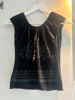 Svart blus med blått mönster och snörning - Cool svart Y2K blus med blått broderat mönster och snörning med träpärlor framtill. Blusen har korta ärmar och ett dekorativt band vid halsen. Perfekt för dig som gillar unika detaljer och boho-vibes. Storlek säger S/M, men den passar mer som XS/S. 