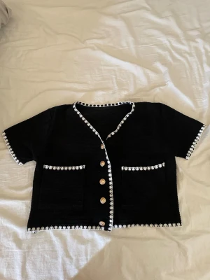 Svart kortärmad kofta med vita detaljer - Svart kortärmad kofta med vita kantdetaljer och dekorativa knappar framtill. Två fickor framtill med vit kant. Tillverkad i en mjuk blandning av viskos, polyester och spandex. Perfekt för en trendig och stilren look.