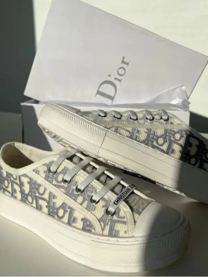 Dam Skor - Säljer ett par Dior B23 låga sneakers med ikoniskt grått och vitt monogrammönster. Skorna har vita snören, robust vit sula och detaljer i canvas. Klassisk Dior-logga på sidan och metallögla med texten 'Christian Dior'. Perfekt för dig som gillar lyxig streetstyle.