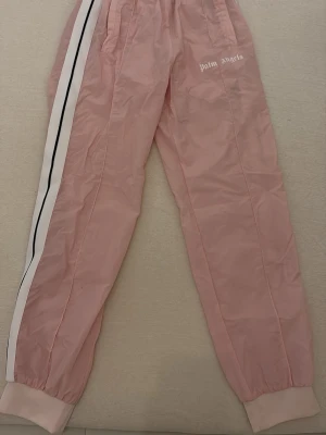 Palm Angels rosa mjukisbyxor - Säljer ett par ljusrosa mjukisbyxor från Palm Angels med vita och svarta sidoremsor. Byxorna har resår i midjan och vid bensluten samt fickor framtill. Materialet är lätt och har en sportig känsla. Perfekta för en chill och trendig look. Priset är ej hugget i sten. Skriv för frågor