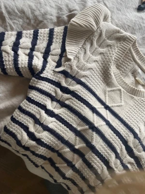 Randig stickad tröja med volang - Krämvit stickad tröja med mörkblå ränder och volangdetaljer vid axlarna. Tröjan har ett snyggt kabelstickat mönster framtill, rund halsringning och ribbade muddar. Perfekt för dig som gillar en trendig och mysig look.