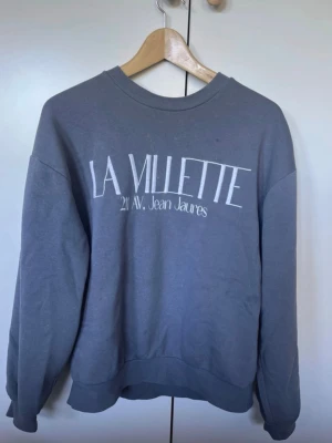Grå sweatshirt LA VILLETTE - Mysig grå sweatshirt med trycket 'LA VILLETTE 211 AV. Jean Jaurès' framtill. Tröjan har rund halsringning och långa ärmar med ribbade muddar. Perfekt för en avslappnad och stilren look.