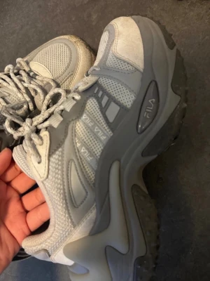 Grå chunky sneakers från Fila - Säljer ett par grå chunky sneakers från Fila med robust sula och meshdetaljer. Skorna har en sportig vibe med blandning av ljusgrått och mörkgrått, samt Fila-logga på sidan och bak. Perfekta för dig som gillar streetwear och vill ha bekväma skor med cool design.