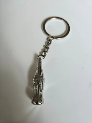 Nyckelring Coca-Cola - Snygg nyckelring med ett hänge i form av en klassisk Coca-Cola läskflaska i silverfärgad metall. Perfekt accessoar för dig som gillar retrostil och vill ha något unikt på nyckelknippan. Metallkedja och ring för enkel fastsättning.