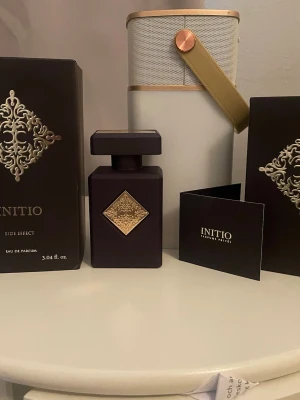 Initio Side Effect Eau de Parfum - Initio Side Effect Eau de Parfum i en lyxig mörklila flaska med guldfärgad dekor på framsidan. Flaskan har en fyrkantig form och matchande lock. Kommer i elegant svart förpackning med gulddetaljer. Doften är exklusiv och passar dig som gillar unika parfymer.
