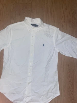 Vit skjorta från Polo Ralph Lauren - Klassisk vit skjorta från Polo Ralph Lauren med blå broderad logga på bröstet. Skjortan har knappar framtill, lång ärm och button-down krage. Tillverkad i bomull för en bekväm och stilren look.