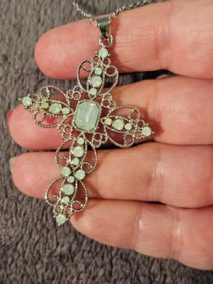 Silverfärgat halsband med korshänge - Snyggt halsband i silverfärg med ett stort korsformat hänge dekorerat med vita pärlor och gnistrande stenar. Hänget har en detaljerad, vintageinspirerad design med många små pärlor och ett större, rektangulärt centrum. Perfekt för dig som gillar statement-smycken.