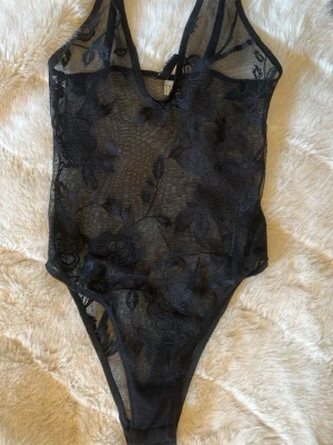 Svart spetsbody med blommönster - Snygg svart body i transparent spets med blommönster. Djup ringning fram och smala axelband. Perfekt för att skapa en trendig och stilren look. Materialet är stretchigt och följsamt för en bekväm passform.