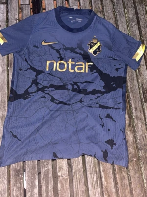 AIK matchtröja Nike blå/guld L - AIK:s officiella matchtröja från Nike i blått med guldiga detaljer och klubbmärke på bröstet. Tröjan har ett unikt kartmönster, Dri-Fit ADV-material och slim fit-passform. Perfekt för fotboll eller träning.