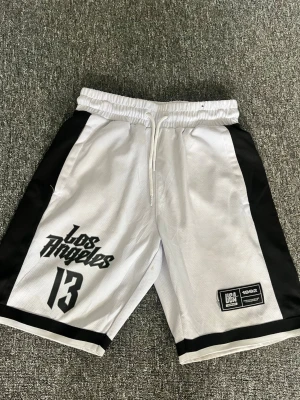 Vita och svarta Los Angeles shorts - Snygga basketinspirerade shorts i vitt med svarta sidopaneler och tryck med texten 'Los Angeles' och siffran 13. Shortsen har elastisk midja med snörning och är tillverkade i ett sportigt, ventilerande polyestermaterial. Den har några fläckar som förmodligen går bort i tvätten
