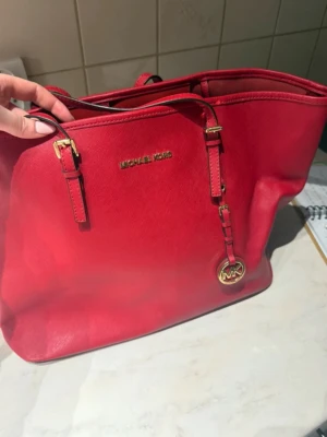 Röd handväska/totebag från Michael Kors - Snygg röd handväska från Michael Kors i slätt läder med guldfärgade detaljer och MK-logga. Väskan har dubbla axelband och en klassisk, rymlig design som passar perfekt för dig som vill ha både stil och funktion.