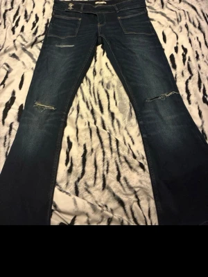 Mörkblå bootcut jeans med slitningar - Säljer ett par mörkblå low waist bootcut jeans med slitningar vid knäna och framsidan. Jeansen har två framfickor och dekorativa sömmar. Perfekta för dig som gillar en avslappnad och trendig stil. De är uppsydda och ni har måttet i slutet💕