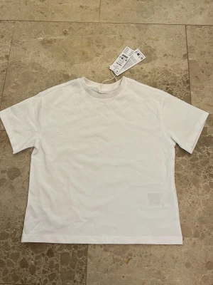 Vit basic t-shirt från Gina Tricot - Klassisk vit t-shirt från Gina Tricot i mjuk bomull. Modellen har rund halsringning och korta ärmar, perfekt för en clean och enkel stil. Passformen är normal och materialet känns skönt mot huden. En riktig garderobsklassiker!