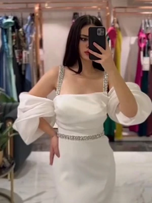 Festklänningar  - Elegant vit långklänning med glittrande axelband och bälte i strass. Klänningen har offshoulder-ärmar med volang och är tillverkad i ett glansigt satinliknande material. Perfekt för bal eller student. Snygg och unik design som verkligen sticker ut.