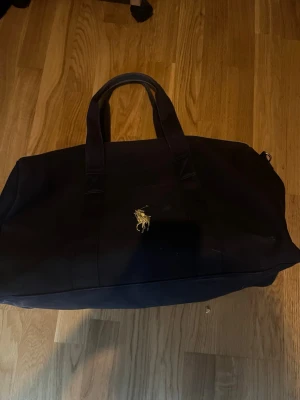Svart weekendbag från Polo Ralph Lauren - Stilren svart weekendbag från Polo Ralph Lauren med klassisk guldfärgad logga framtill. Väskan har dubbla handtag och rymligt huvudfack med dragkedja. Perfekt för resor eller gymmet. Tillverkad i slitstarkt tygmaterial.