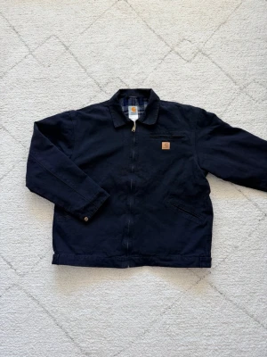 Marinblå Carhartt jacka med rutigt foder - Säljer en marinblå Carhartt jacka med dragkedja och klassisk krage. Jackan har ett rutigt foder i blått, vitt och beige samt två snedställda fickor framtill och en bröstficka. Carhartt-logga på bröstet. Perfekt för dig som gillar streetwear och robust stil.