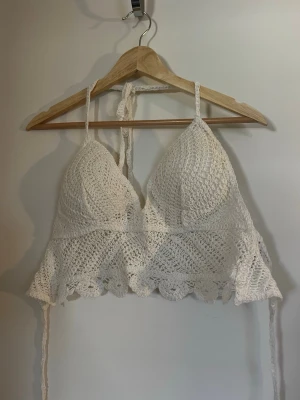 Vit virkad topp med knyt - Supersöt virkad linnetopp i vitt med knyt i nacken och ryggen. Toppen har en scallop-kant nertill och är ärmlös, perfekt för sommardagar. Detaljerad virkning ger en bohemisk känsla och toppen är lätt att styla till olika outfits.