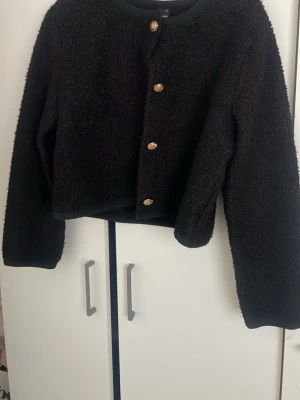 Svart teddykofta från Gina Tricot - Mysig svart teddykofta från Gina Tricot med fyra dekorativa guldfärgade knappar framtill. Koftan har rund halsringning och är tillverkad i ett fluffigt, mjukt material som ger en trendig look. Perfekt för lager på lager och enkel att styla till olika outfits.