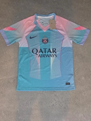 PSG matchtröja i pastell från Nike - Säljer en PSG matchtröja från Nike i snygga pastellfärger med blå, rosa och vita toner. Tröjan har korta ärmar, V-ringning och tryck med Qatar Airways på bröstet. Perfekt för dig som älskar fotboll och vill sticka ut på planen.