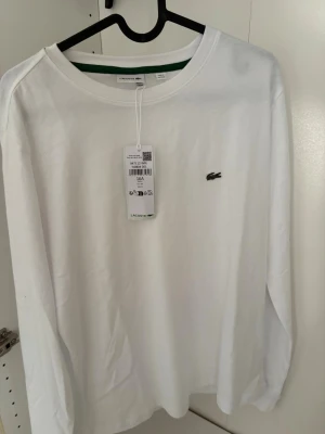 Vit långärmad topp från Lacoste - Klassisk vit långärmad topp från Lacoste med rund halsringning och den ikoniska gröna krokodilloggan broderad på bröstet. Tillverkad i mjuk bomull för en clean och stilren look. Perfekt till jeans eller kjol.