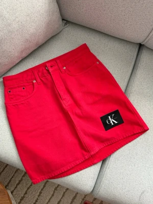 Röd kortkjol från Calvin Klein - Snygg röd kortkjol från Calvin Klein med klassisk femficksdesign och svart CK-logga framtill. Kjolen är i ett stadigt bomullsmaterial och har bälteshällor samt knapp- och dragkedjestängning. Perfekt för dig som vill sticka ut med färg!