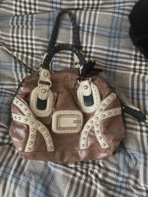 Brun och beige handväska från Guess - Säljer en cool handväska från Guess i brunt och beige konstläder med nitar och dekorativa detaljer. Väskan har flätade handtag i svart och ett stort Guess-emblem framtill. Perfekt för dig som gillar statement-accessoarer. Vintage och verkar inte skapas längre!