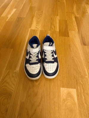 Nike Dunk Low blåvita sneakers - Snygga Nike Dunk Low sneakers i blått och vitt med klassisk swoosh på sidan. Skorna har snörning och kardborreband upptill för extra support. Ovandelen är i läder och mesh, vilket ger både stil och komfort. Perfekta för dig som gillar streetwear och sportig look.