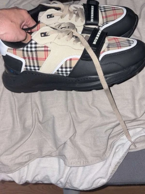 Burberry sneakers med rutigt mönster - Snygga sneakers från Burberry med klassiskt beige, svart och rött rutmönster, svarta och vita detaljer samt beige mockaliknande partier. Skorna har bred svart sula och Burberry-logga på häl och kardborreband. Perfekta för dig som vill sticka ut med lyxig streetstyle.