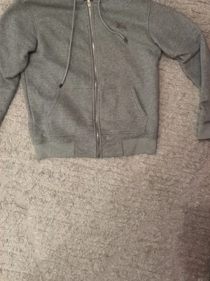 Grå zip-hoodie från Burberry - Grå hoodie med dragkedja från Burberry. Klassisk modell med huva och dragsko, två fickor framtill och diskret Burberry-logga på bröstet. Tillverkad i mjuk bomull och fodrad med det ikoniska Burberry-mönstret på insidan av huvan. Skick 10/10 använt endast fåtal gånger. Storlek M men passar även S