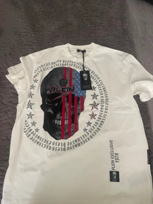 Vit t-shirt med dödskalle från Plein - Säljer en vit t-shirt från Plein Exclusive med stor dödskalle och amerikanskt flaggmotiv framtill. T-shirten har korta ärmar, tryckta stjärnor och text runt motivet. Tillverkad i 100% polyester och har en cool streetwear vibe.