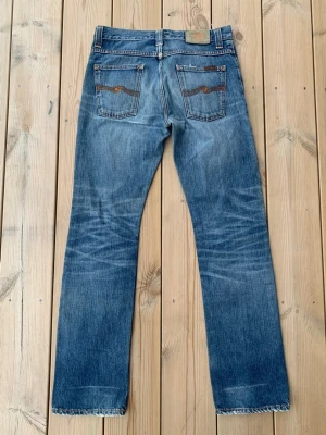 Nudie Jeans Average Joe - Säljer ett par ljusblå jeans med tydliga slitningar och hål på låren och knäna. Jeansen har rak passform och klassisk femficksdesign. Perfekta för en avslappnad och trendig look. Materialet är denim i bomull.