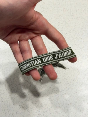 Grönt armband - Snyggt vävt armband med text i vitt på en olivgrön bakgrund. Armbandet har justerbara tofsar i ändarna och är gjort av textilmaterial. Perfekt accessoar för att lyfta din outfit med en exklusiv touch.